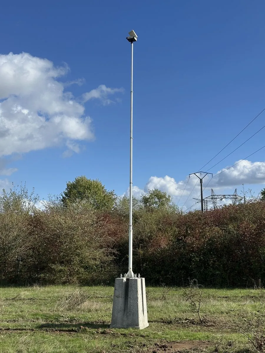 Mât télescopique blanc fixé sur un bloc béton au milieu d’un terrain nu, la caméra timelapse en haut, des pylônes haute tension au loin