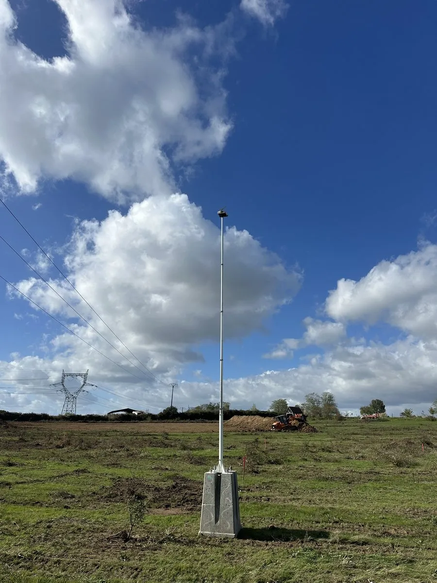 Le mât télescopique sur son bloc béton, seul au milieu d’un vaste terrain enherbé, sous un ciel nuageux, des pylônes haute tension à l’horizon